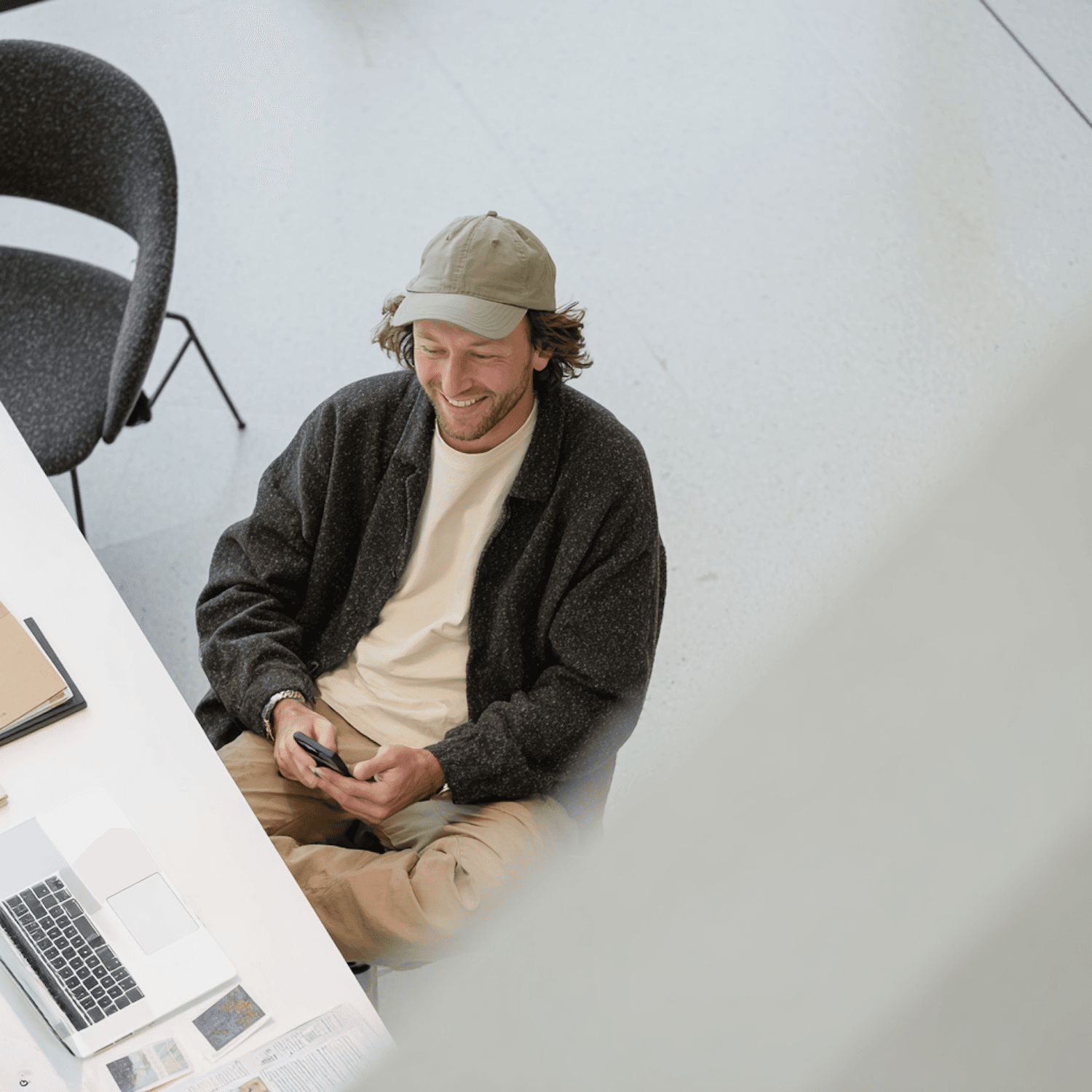 Homem sentado à mesa de trabalho olhando o celular, com notebook e papéis ao lado